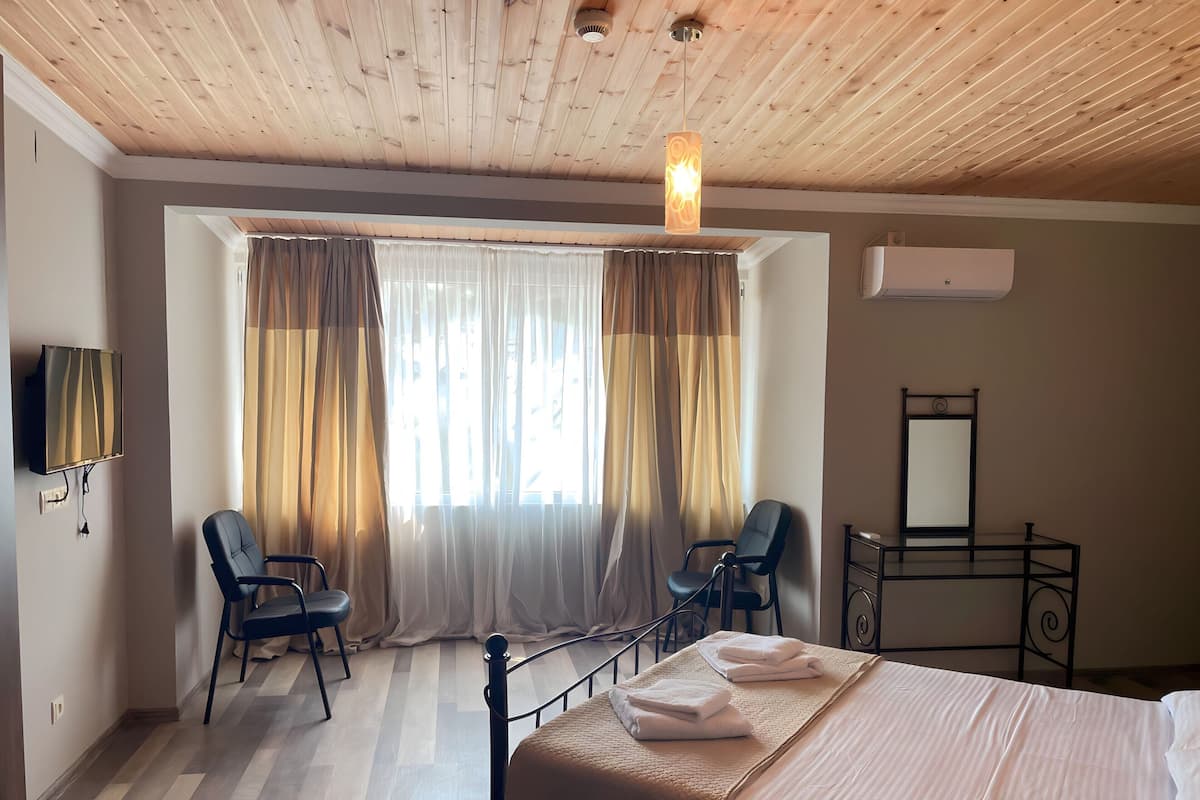 A hotel room with a wooden ceiling, a bed with a white bedspread, two chairs, a small table, a TV mounted on the wall, and a window with curtains.