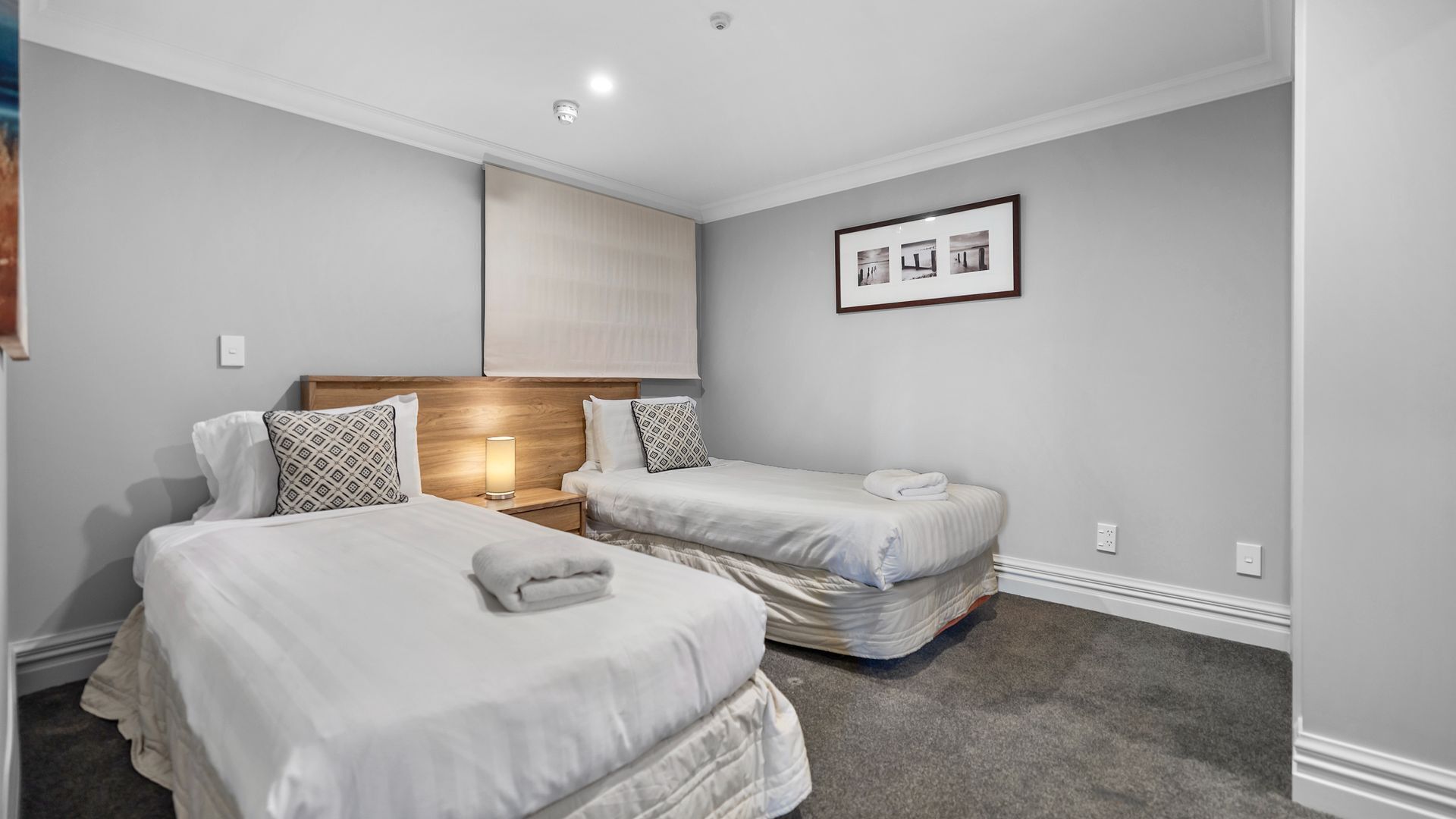 Habitación de hotel con dos camas, un cabecero de madera, una mesita de noche con lámpara y un cuadro en la pared.