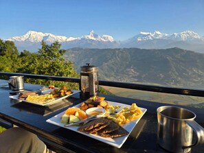 A meal set on a table with a mountain view, including a plate with toast, scrambled eggs, and fruit, a thermos, and a metal cup.
