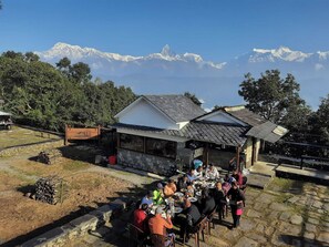 A dining area with tables and chairs, a stone building with a tiled roof, and a mountainous backdrop.
