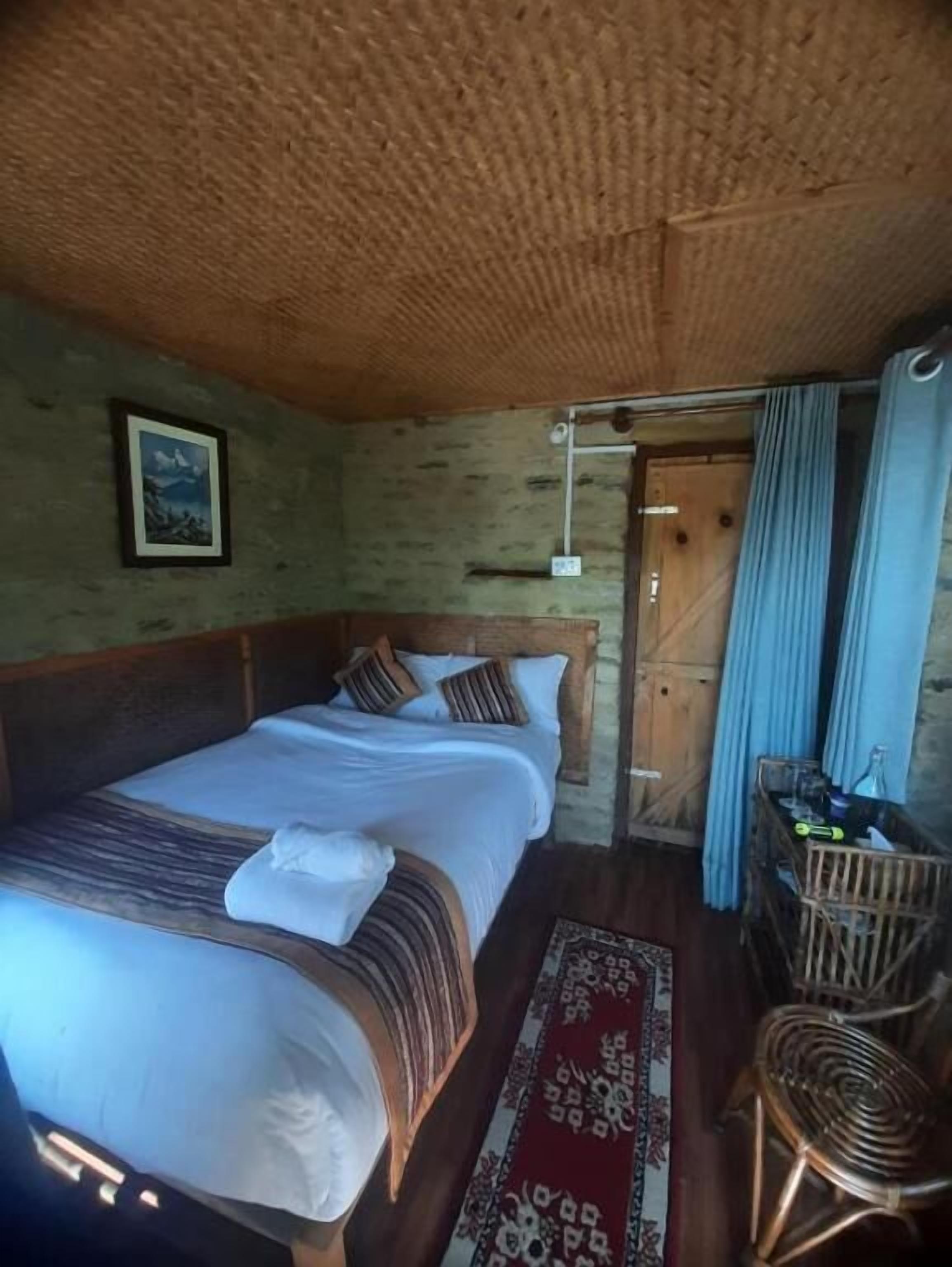 A room with two beds, a wooden ceiling, a framed picture on the wall, a wooden door, and a small table with items.