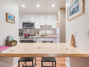 Cozinha compacta com armários brancos, micro-ondas de aço inoxidável e mesa de madeira com dois bancos.