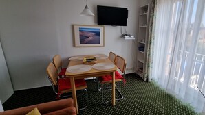 Ein Zimmer mit Esstisch, Stühlen, einem an der Wand befestigten Fernseher, einem Bücherregal und einem Fenster mit Vorhängen.