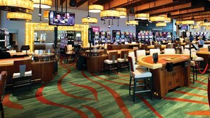 A casino with multiple gaming tables and chairs, a bar area, and a large screen displaying a game.
