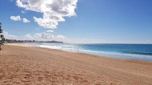 Una playa extensa de arena clara con olas suaves, cielo despejado y colinas lejanas.