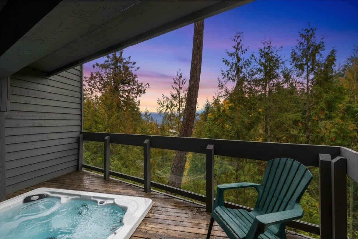 A wooden deck with a hot tub and a green Adirondack chair, overlooking a forested area.