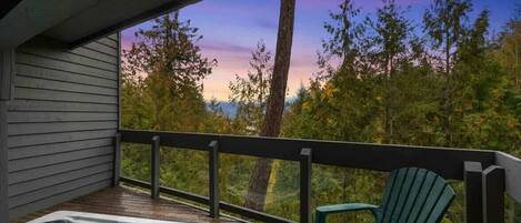 A wooden deck with a hot tub and a green Adirondack chair, overlooking a forested area.