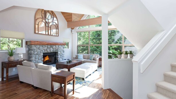 Amplia sala de estar con chimenea de piedra, vigas de madera y grandes ventanales con vistas a un bosque.