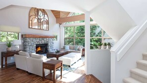 A spacious living room with a stone fireplace, wooden beams, and large windows overlooking a forest. - Cozy townhouse w/ hot tub & ski-access (Whistler)