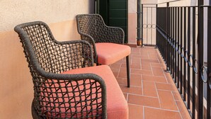 Un balcón con dos sillas de mimbre y un cojín rojo, piso de baldosas y baranda de metal negro.