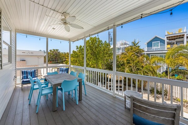 A covered outdoor deck with a table and chairs, a ceiling fan, and a view of neighboring buildings and trees.