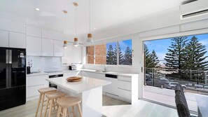 A modern kitchen with white cabinetry, a central island with stools, a large window with a view of trees and a snowy landscape, and a black refrigerator. - Blue Pacific Apartment 8 - Fully renovated 3 bedroom on marine Parade. (The Entrance)