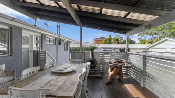 A covered outdoor patio with a wooden table and chairs, a stone fire pit, and a chimney.