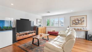 A living room with a flat-screen TV, wooden entertainment unit, red sofa, beige sofa, and a large window with blinds.