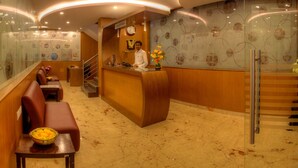 A hotel reception area with a receptionist, a reception desk, seating area, and a staircase. - Hotel Aditya Mysore (Mysore)
