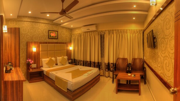 A hotel room with a large bed, a desk, two chairs, a ceiling fan, and a TV mounted on the wall. - Hotel Aditya Mysore (Mysore)