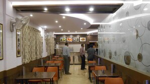 A restaurant with a long counter, orange chairs, and a glass wall displaying food items. - Hotel Aditya Mysore (Mysore)