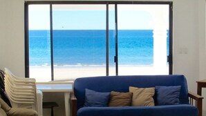 Quarto com uma grande janela que oferece vista para o oceano. Há um sofá azul com almofadas e um sofá bege.