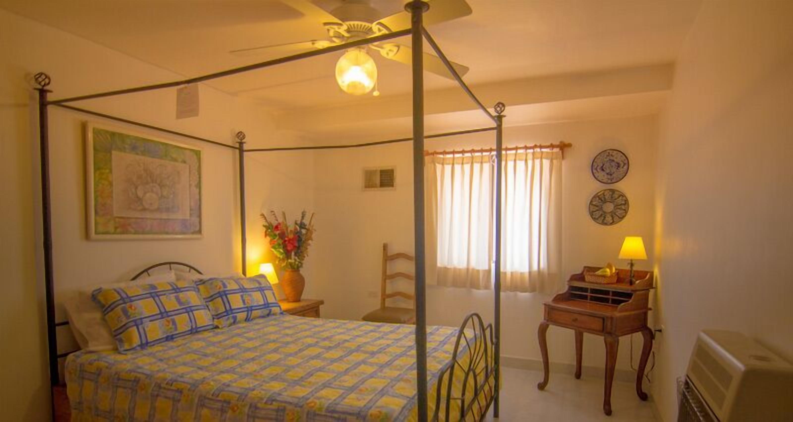 A four-poster bed with a patterned bedspread, a bedside table with a lamp, a chair, and a wall clock.