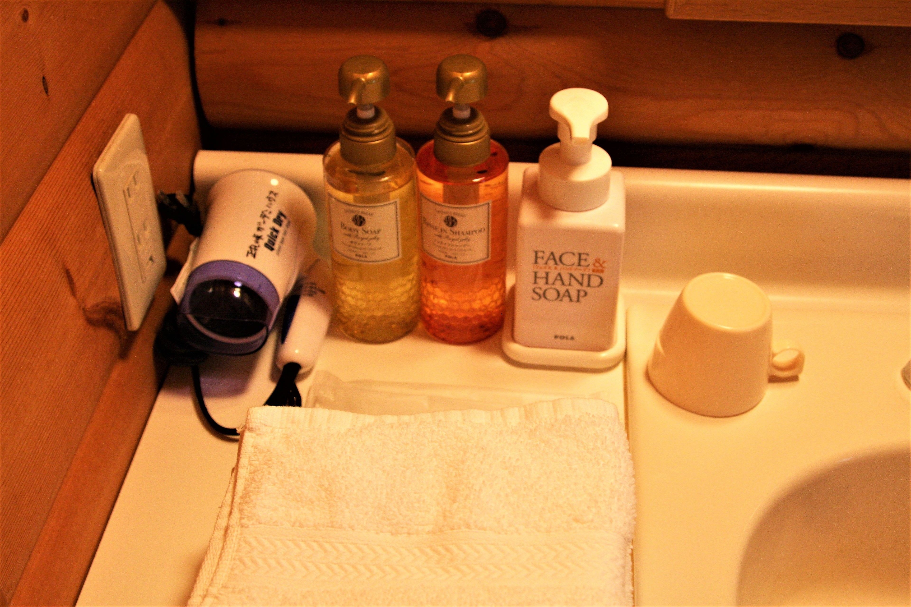 A bathroom sink area with a white towel, two bottles of shampoo, a bottle of face and hand soap, and a white cup.