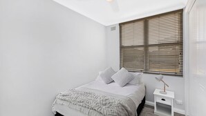 A bedroom with a bed, pillows, a bedside table, and a window with blinds. - Golden Sands Apartment 10 (Blue Bay)