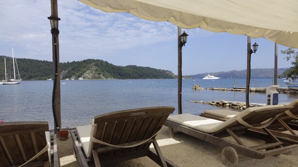 A beachside lounge with wooden chairs and a canopy, overlooking a calm sea with boats and a hilly landscape in the distance. - Sakin Vadi Winehouse (Marmaris)