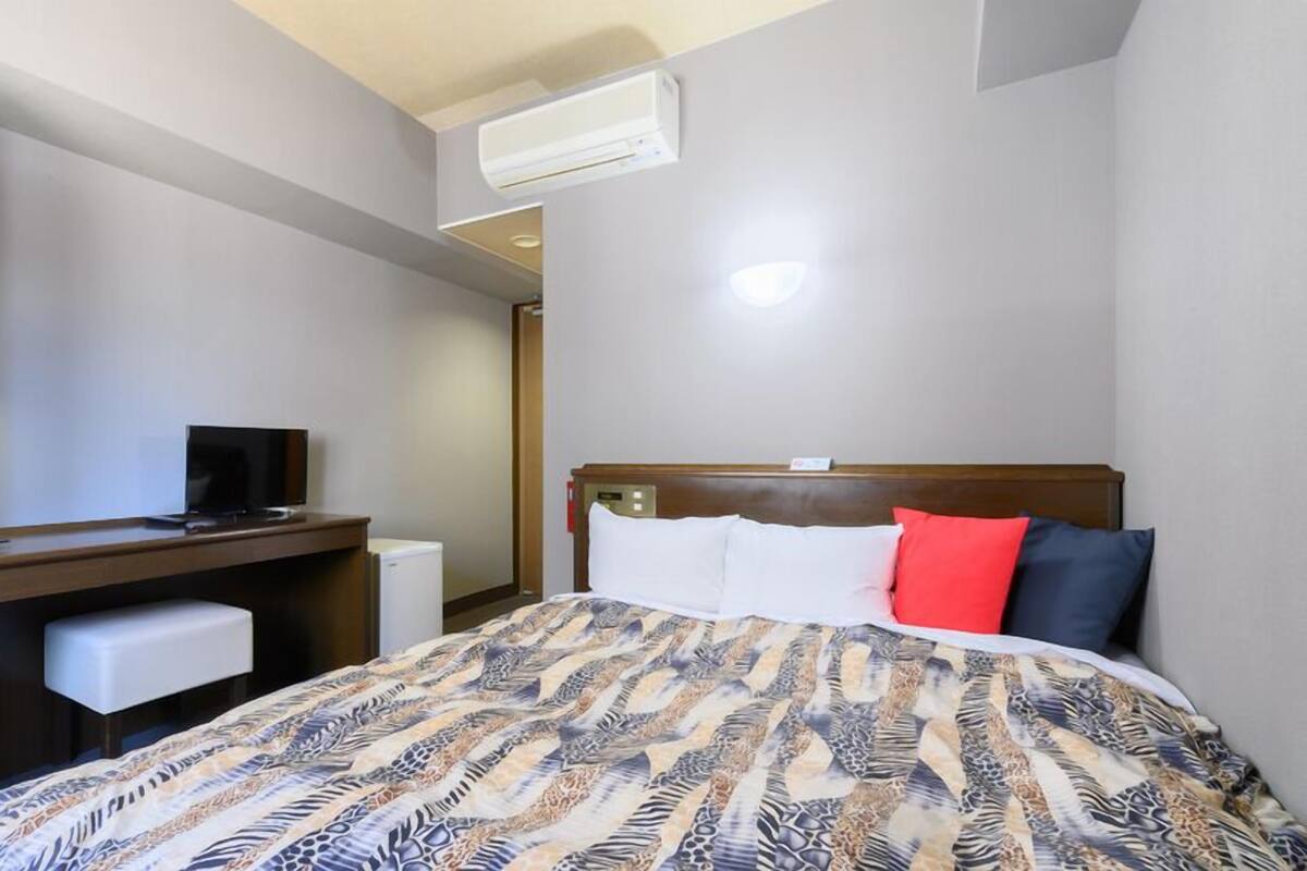 A hotel room with a bed, a desk, a television, and an air conditioning unit.