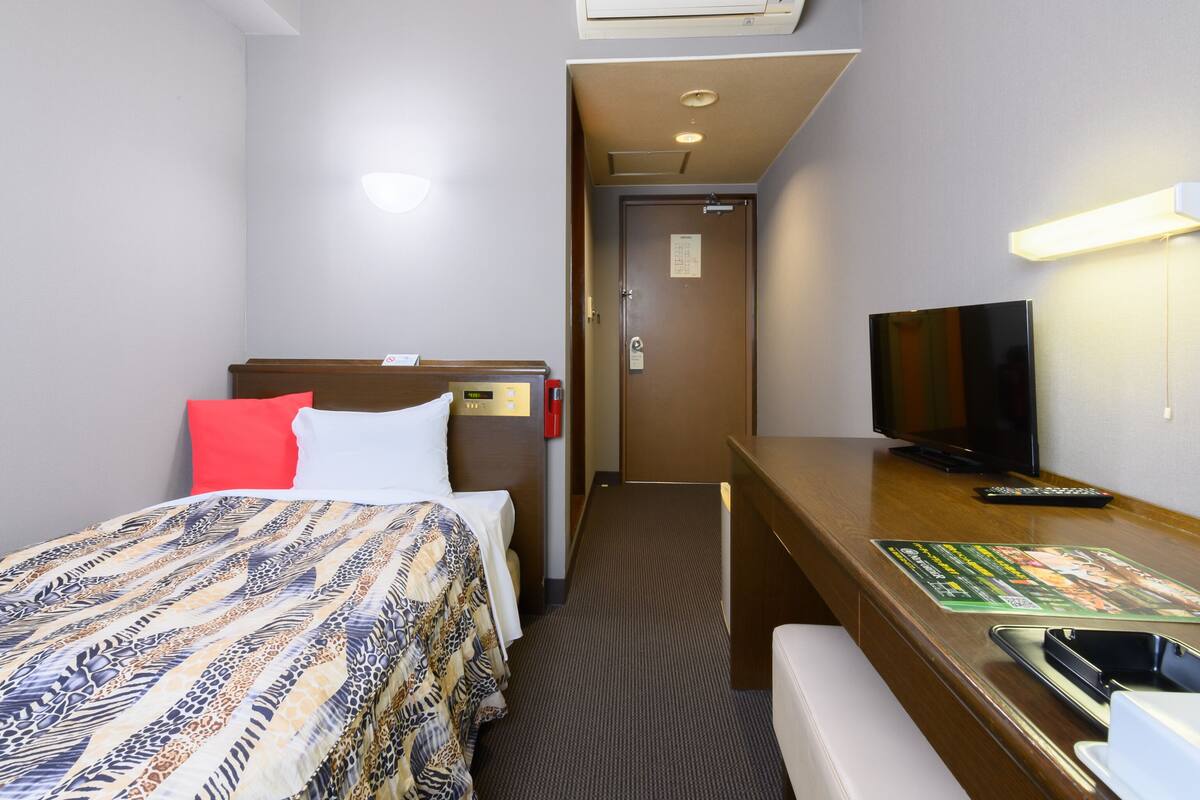 A hotel room with a bed, a desk with a television, a chair, a small table, and an air conditioning unit.