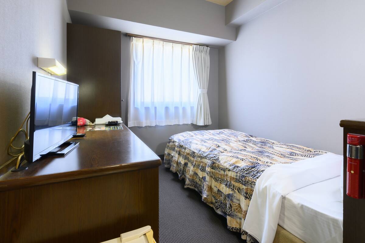 A hotel room with a bed, a desk with a television, a window with curtains, and a red fire extinguisher.