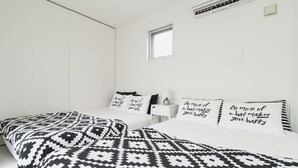A modern bedroom with a large bed, a small bedside table, a nightstand, a window, and an air conditioning unit. - Niraikanai (Nanjo)