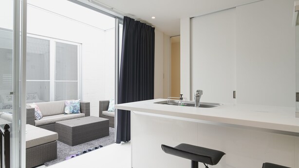 A modern kitchen with a white countertop, a sink, and a bar stool. There is a dining area with a sofa and a small table. - Niraikanai (Nanjo)