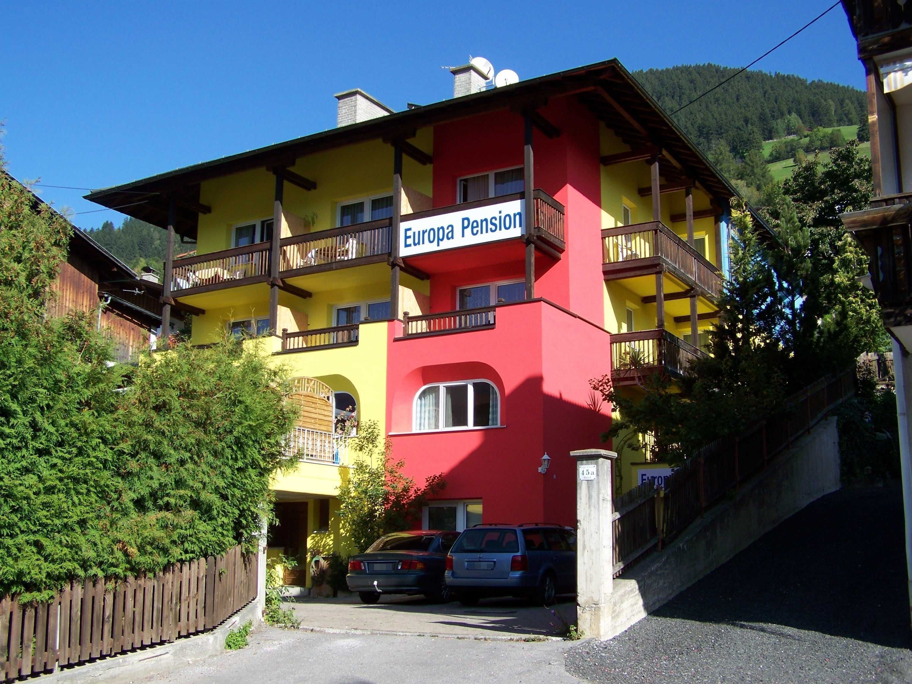A multi-story building with balconies, labeled 'Europa Pension,' featuring a red and yellow facade, parked cars, and a mountainous backdrop.