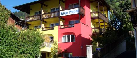 A multi-story building with balconies, labeled 'Europa Pension,' featuring a red and yellow facade, parked cars, and a mountainous backdrop.