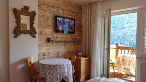 Une pièce avec un mur en bois, un téléviseur, une table recouverte d’une nappe blanche, une chaise en bois, un buffet en bois et une vue sur une forêt, visible à travers une porte vitrée.