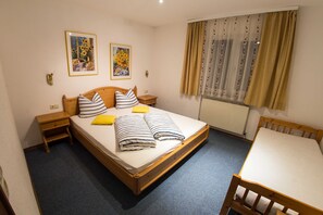 Una cama individual con estructura de madera, dos mesitas de noche, una pequeña mesa y una ventana con cortinas.