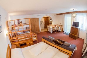 Ein Zimmer mit einem Bett, einem Holzbodenregal, einem Esstisch mit Stühlen, einem Fernseher und einer Küchenzeile.