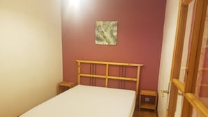 A single bed with a wooden frame and a white mattress in a room with a red wall and a small painting. - ---- Villa Lez ---- (Prades-le-Lez)