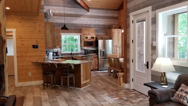 A cabin interior with a wooden interior, a kitchen with a central island, bar stools, a dining table, and a couch with a 'Cabin' pillow. - Storybook Caretaker's Cabin in the Woods -Clemson (Central)
