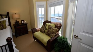Une chambre à coucher comprenant un lit, une table de chevet, un fauteuil en osier donnant sur la mer et une porte.