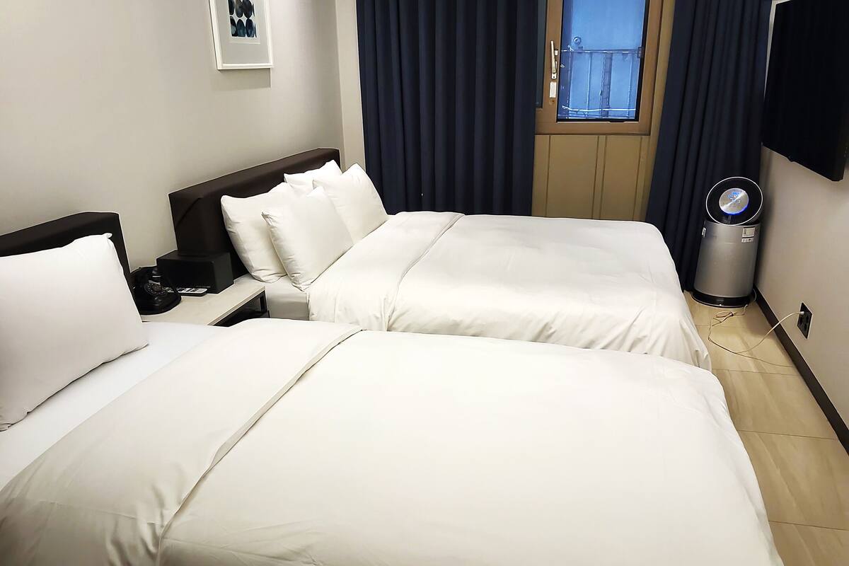 A hotel room with two beds, a bedside table, a lamp, a trash can, and a window with curtains.