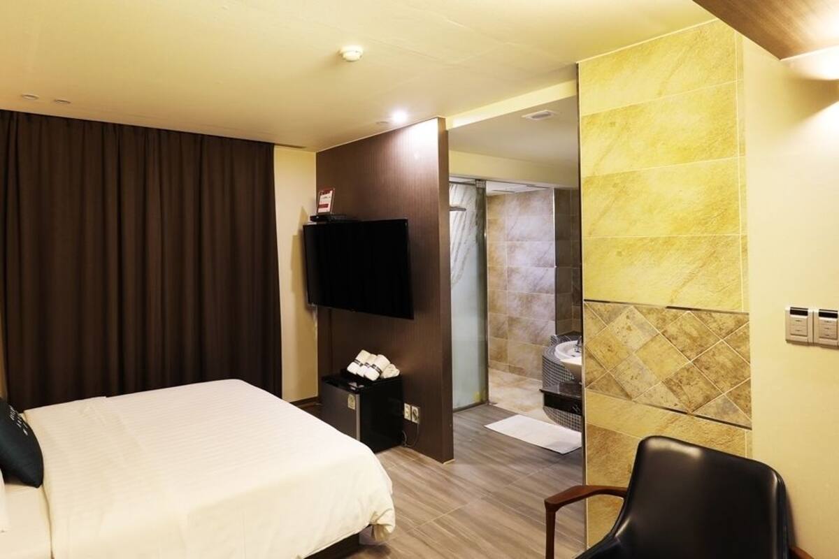 A hotel room with a bed, a TV, a desk, and a bathroom visible through an open door.