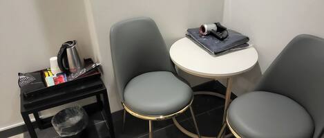 A small room with two grey chairs, a round table, a black side table with a kettle and cups, and a trash bin.