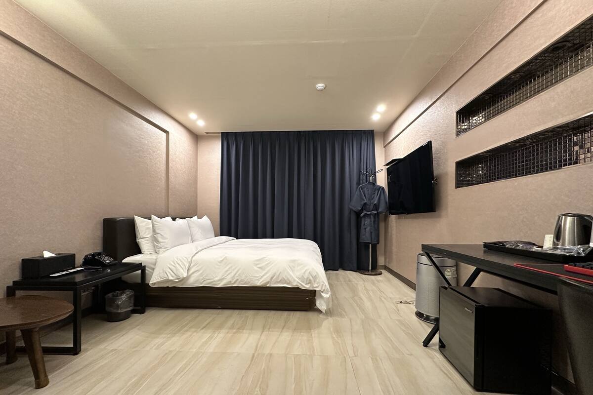 A compact hotel room with a bed, bedside table, a desk with a chair, a TV mounted on the wall, and a wardrobe.