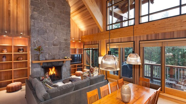 A cozy living room with a stone fireplace, wooden shelves, and a large window overlooking a deck. - Mountain townhouse w/ ski access & hot tub (Whistler)