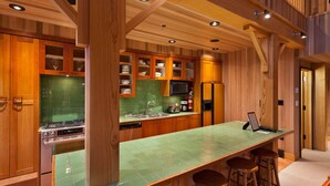 A modern kitchen with wooden cabinetry, a green-tiled backsplash, stainless steel appliances, and a bar area with stools. - Mountain townhouse w/ ski access & hot tub (Whistler)