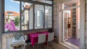 A room with a table and chairs, a window with a view of buildings and flowers, and a door leading to another room.