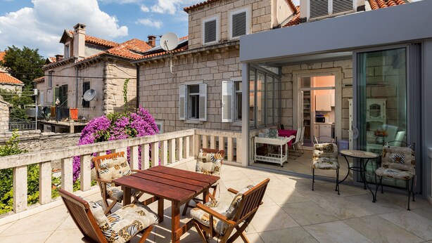 A patio with a wooden table and chairs, a glass door leading to a room, and a balcony with a view of the surrounding buildings.