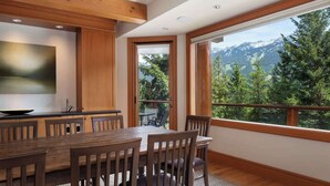Un coin repas avec une table et des chaises en bois, un espace cuisine doté d’un plan de travail noir et une grande fenêtre offrant une vue sur les montagnes.