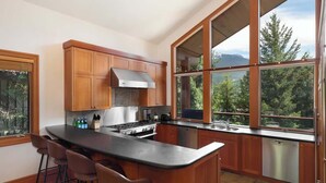 Une cuisine moderne avec des meubles en bois, un îlot central doté de tabourets de bar, une cuisinière en acier inoxydable et de grandes fenêtres offrant une vue sur l’extérieur.