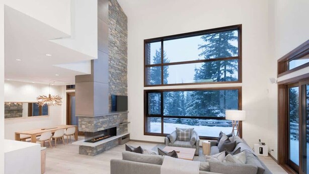 A modern living room with a stone fireplace, large windows with a snowy view, and a cozy seating area. - Lakeside townhouse w/ private garage (Whistler)